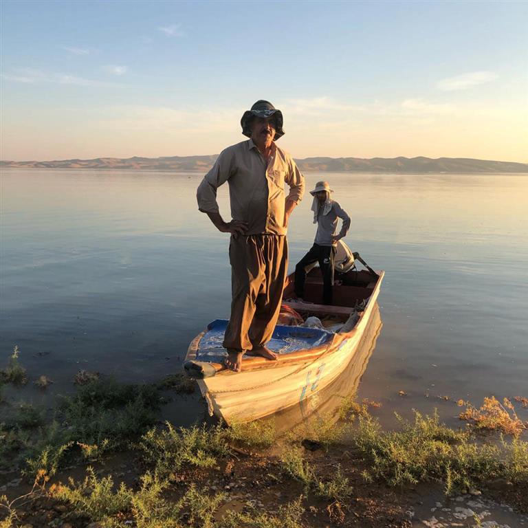 Two Kurdish fishermen standing in a boat on shoals of a body of water.